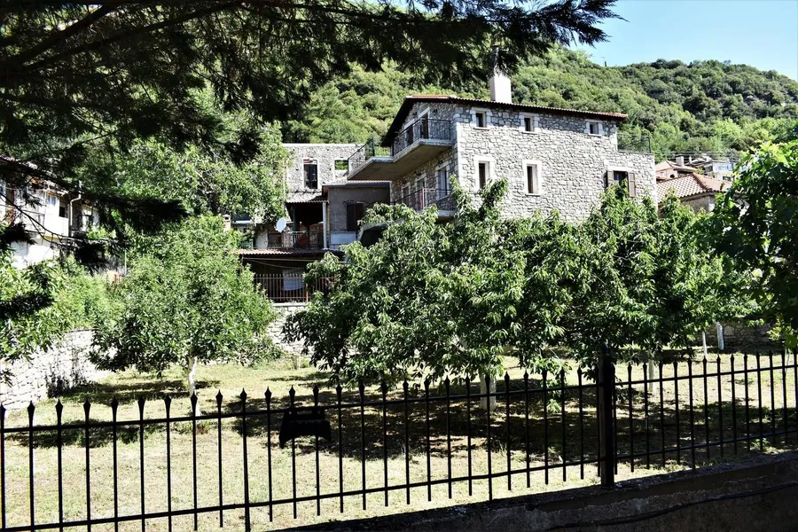 Ο κήπος με τα δέντρα και το πέτρινο σπίτι πίσω από τα κάγκελα
